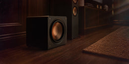 Klipsch Reference Premiere compact sealed subwoofer standing on the floor beside a media console and floorstanding speakers