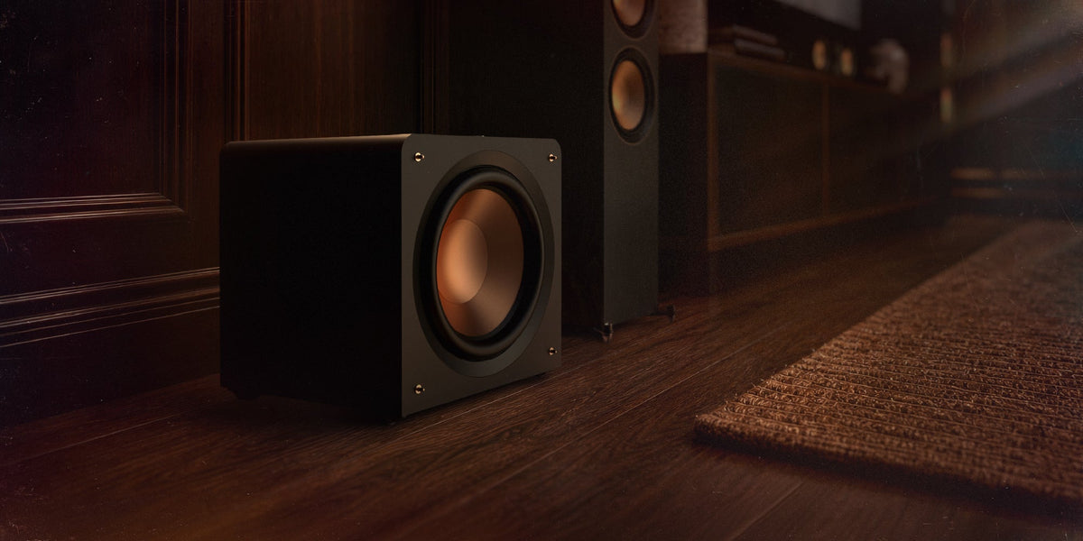 Klipsch Reference Premiere compact sealed subwoofer standing on the floor beside a media console and floorstanding speakers