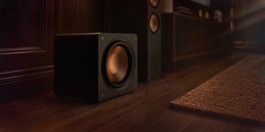 Klipsch Reference Premiere compact sealed subwoofer standing on the floor beside a media console and floorstanding speakers